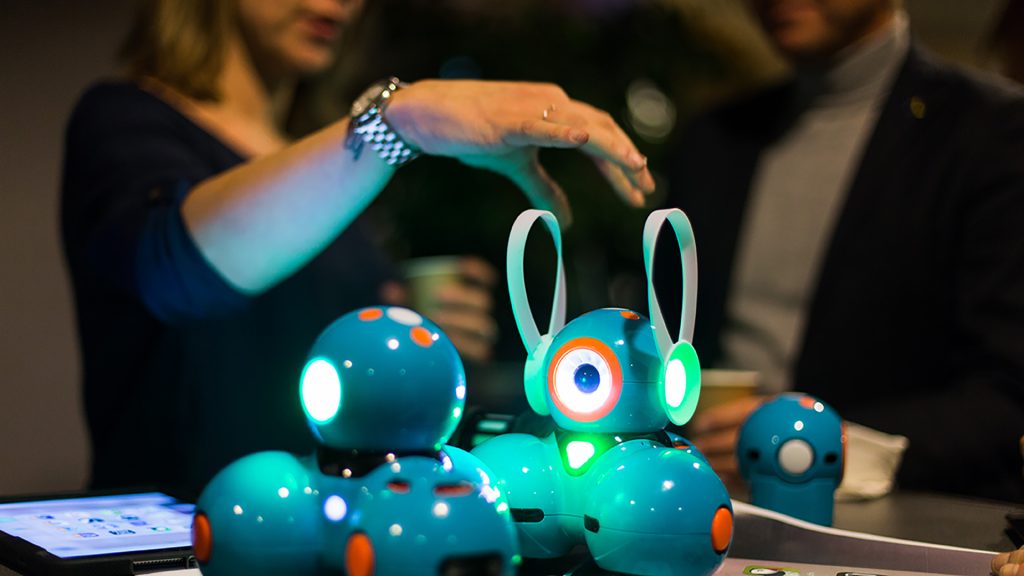 Två robotar som lyser i fokus, bakom står två personer i oskärpa