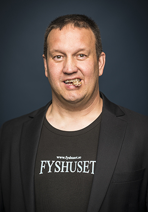 Porträttfoto i studio på Anders Bergström i svart t-shirt med texten Fryshuset på och har ett litet föremål mellan tänderna.