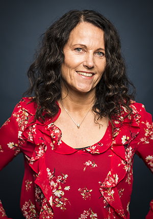 Porträttfoto i studio på Åsa Kock i rödblommig blus och långt lockigt hår i snedbena. 