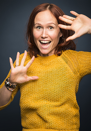 Porträttfoto i studio på Elin Bogren i en orange stickade tröja med båda händerna vända mot kameran.