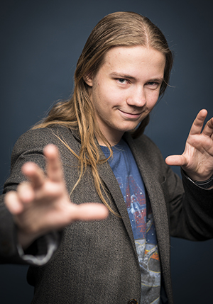 Porträttfoto i studio på Gabriel Larsson i långt hår snett vänd mot kameran och med båda händerna upp i luften.