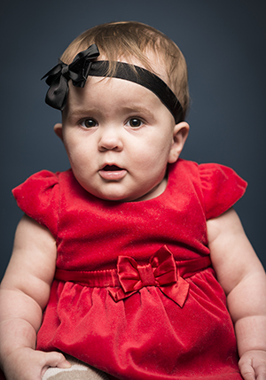 Porträttfoto i studo på Hanna Nordkvist, liten sittande bäbis i röd klänning och svart rosettband i håret.