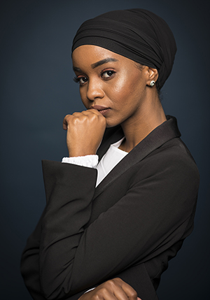 Porträttfoto i studio på Ikram Yusuf med en svart huvudbonad på huvudet och knuten hand hakan.