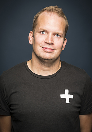 Porträttfoto i studio på Joel Svensson i svart t-shirt med ett vitt kryss på bröstet, ler mot kameran.