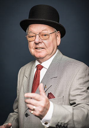 Porträttfoto i studio på Lars Sigvard i en svart hatt, beige kavaj och orange slips med ena handen framför sig.