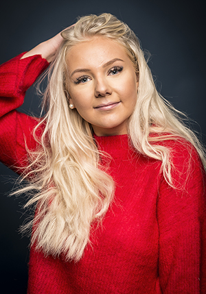 Porträttfoto i studio på Linn Ahlborg i röd stickad tröja och ljust långt hår med ena handen på huvudet.