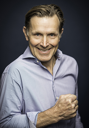 Porträttfoto i studio på Melker Andersson i grå skjorta med knyten näve leendes mot kameran.