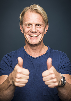 Porträttfoto i studio på Niklas Wikegård i mörkblå t-shirt med båda tummarna upp mot kameran.