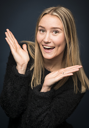 Porträttfoto i studio på Sara McManus gör en sprallig pose med ett brett leende mot kameran.