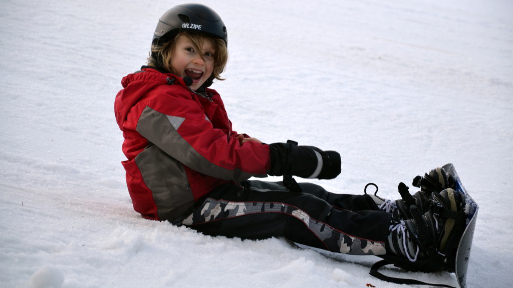 Liten kille sittandes i snö med en snowboard på