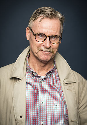 Porträttfoto i studio på Stisse Åberg, en äldre man i beige jacka, rutig skjorta och glasögon.