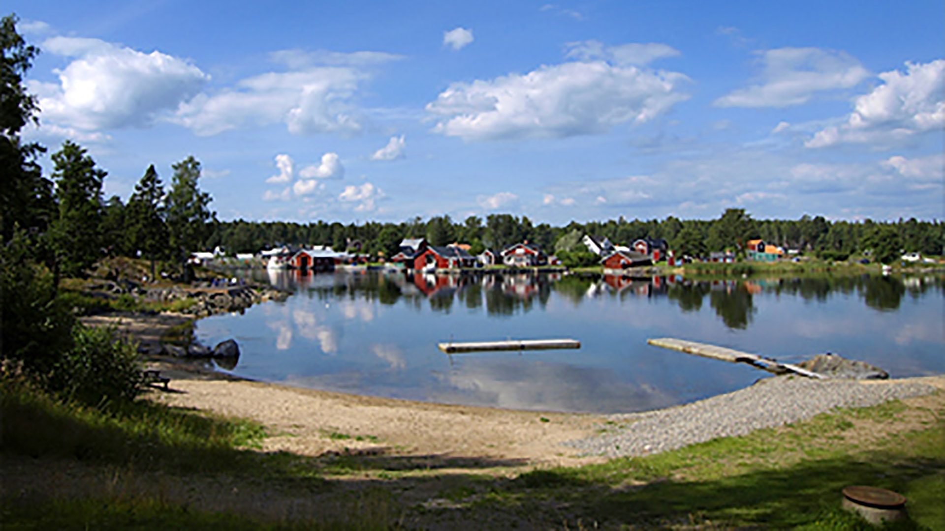 Vatten, badbrygga, sandstrand och gräsmatta vid Saltharsfjärden