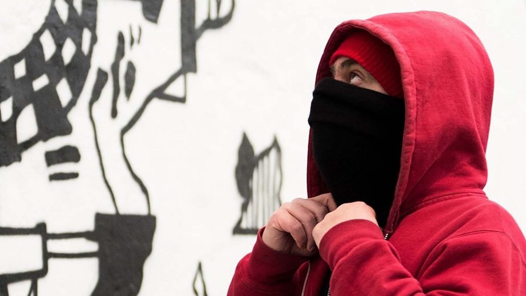 En kille med en röd huvtröja med huvan uppe och ansiktet bakom en scarf. Bara ögonen syns