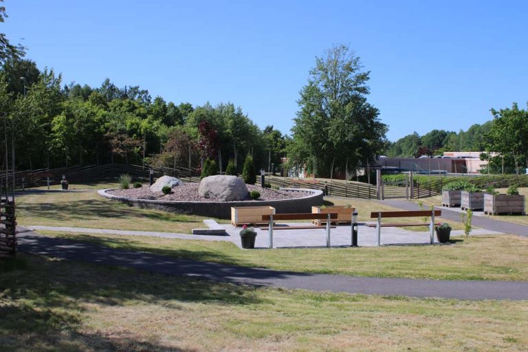 Utomhusmiljö med gröna ytor, odlingskragar och parkbänkar.