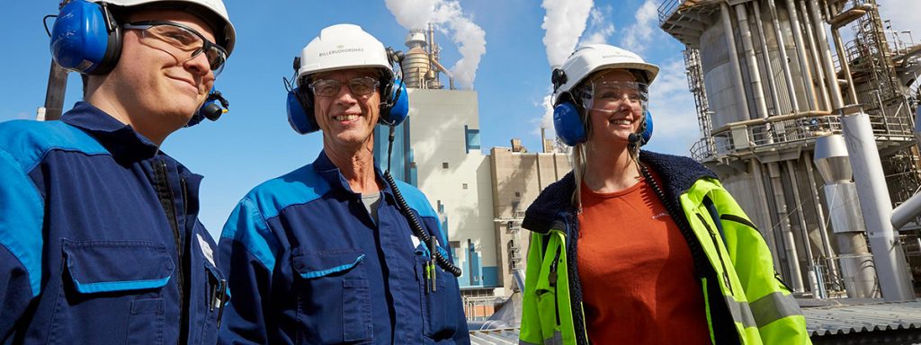 Tre personer med hjälm och hörselkåpor framför en fabrik.