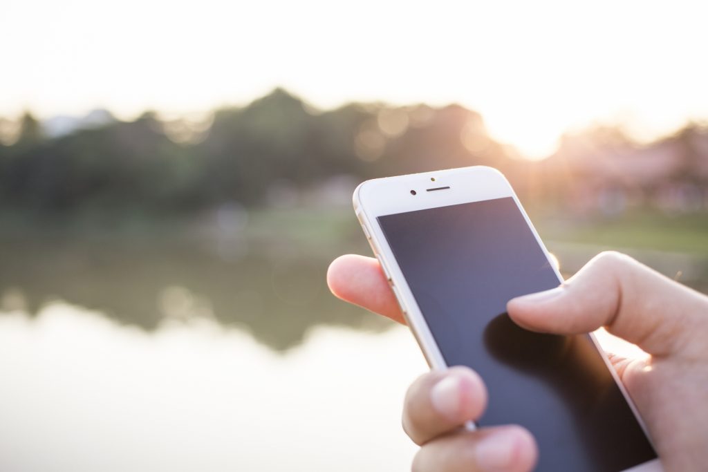 En hand som håller i en vit Iphone med släckt skärm och ett landskap i oskärpa i bakgrunden