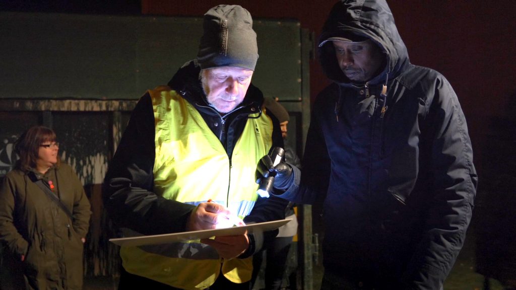 Två män som står i mörker var av en med varselväst och den andra lyser upp med ficklampa
