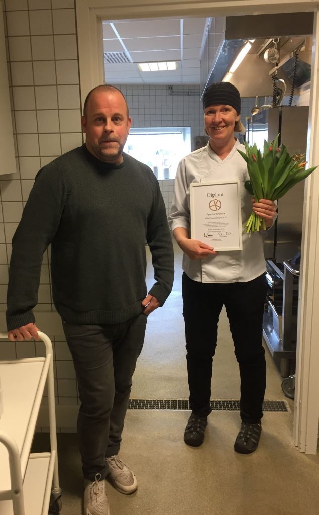En man och kvinna stående i ett restaurangkök. Kvinnan står i kockkläder och håller i ett diplom och bukett.