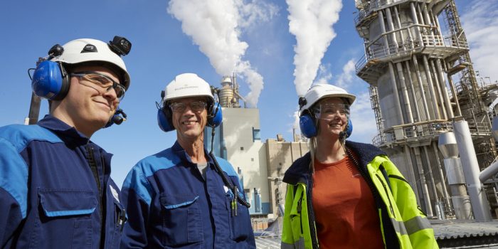 Tre personer med hjälm och hörselkåpor framför en fabrik.