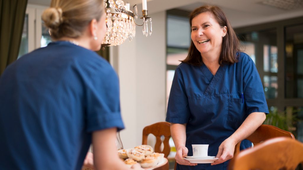 Två kvinnor som arbetar på ett äldreboende och plockar fram fika på ett bord.
