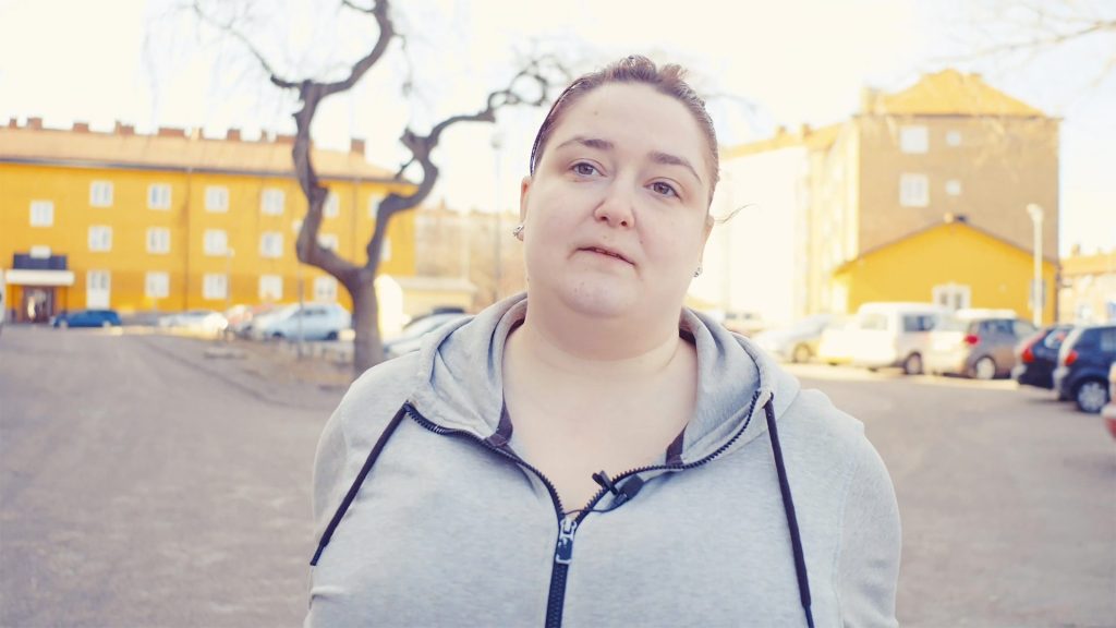 Kvinna i grå munktröja står framförgula hus och ser allvarlig ut.