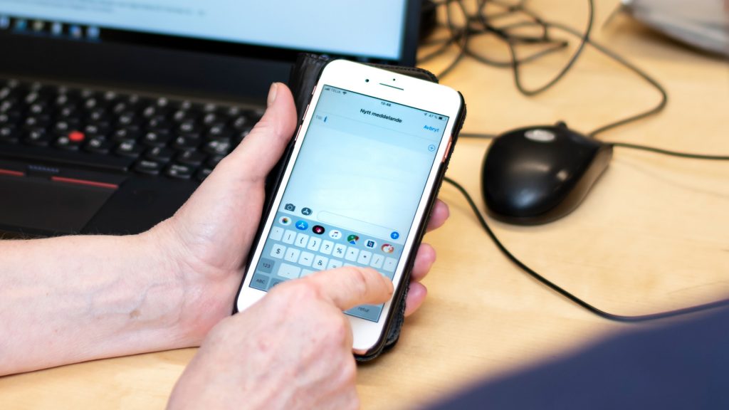Mobiltelefon hålls upp framför en dator där en person ska skriva ett nytt meddelande på telefonen.