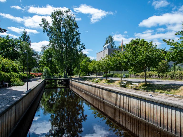 Spegelblank å med bänkar och grönskande träd på sidorna.