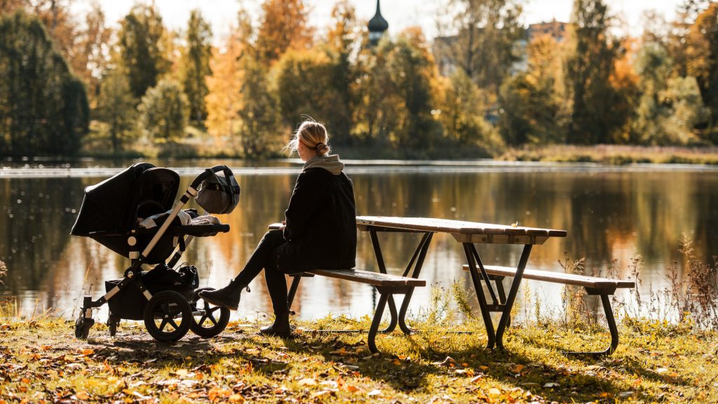 Kvinna med barnvagn som sitter på en parkbänk vid ån.