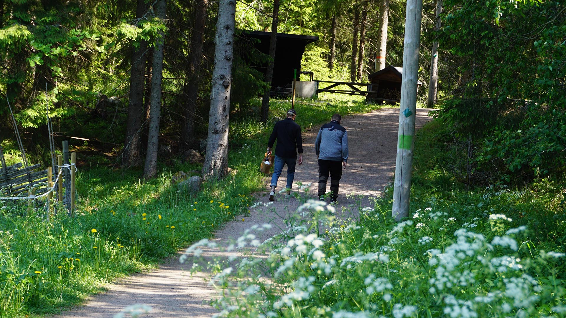 Två personer går i ett motionsspår i skogen. Framför dem på vänster sida finns en grillplats med vindskydd