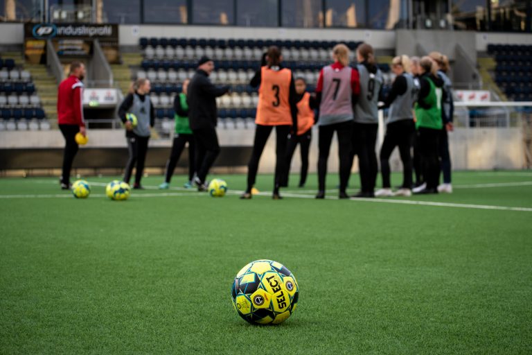 En fotboll i förgrunden, elever på fotbollsplan i bakgrunden