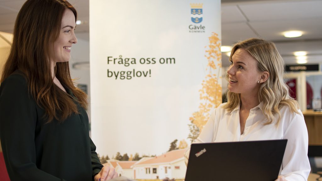 Två kvinnor stående framför en roll up med texten Fråga oss om bygglov!