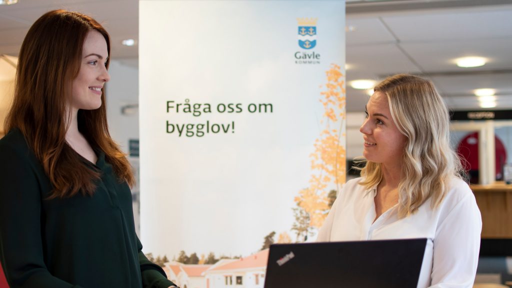 Två kvinnor står framför en roll upp med texten Fråga oss om bygglov.