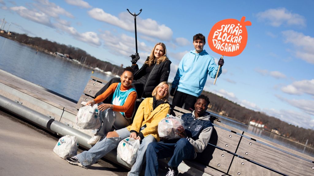 Fem personer som sitter på en bänk vid vatten med skräppåsar