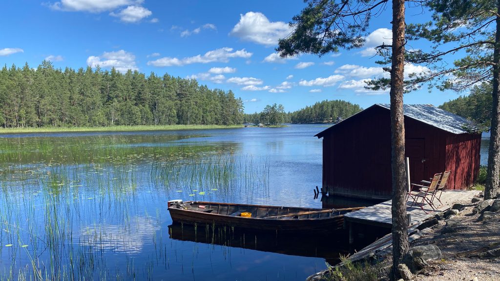 Litet rött hus och eka vid sjö.