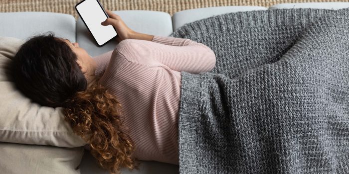 Kvinna ligger i en soffa under en filt och kollar på telefonen