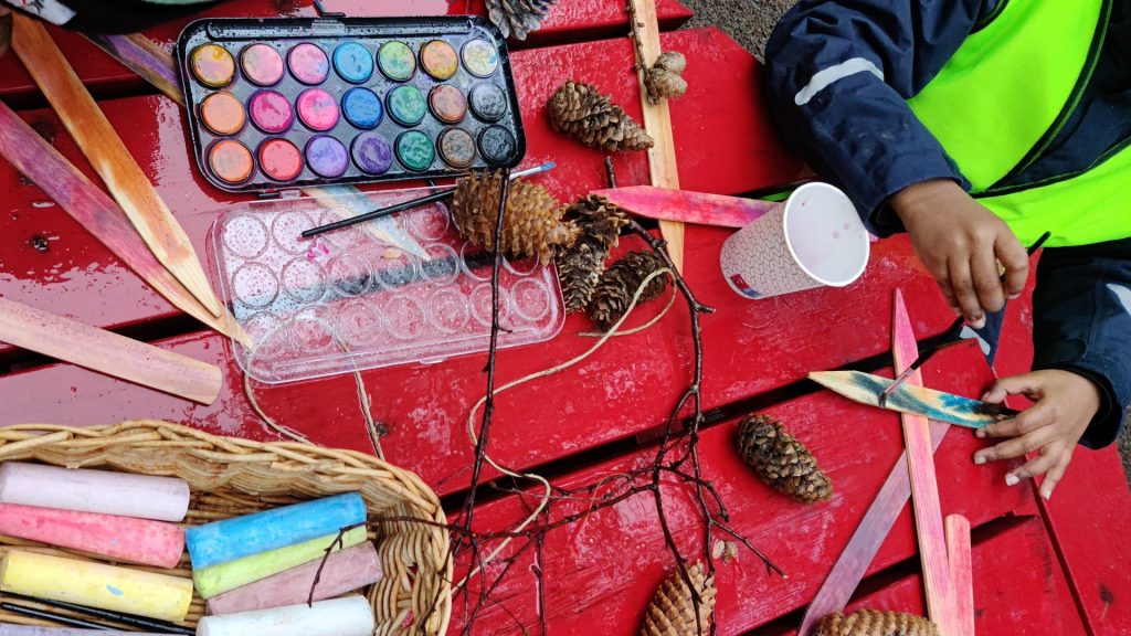 Två händer pysslar med färg, kottar och pinnar