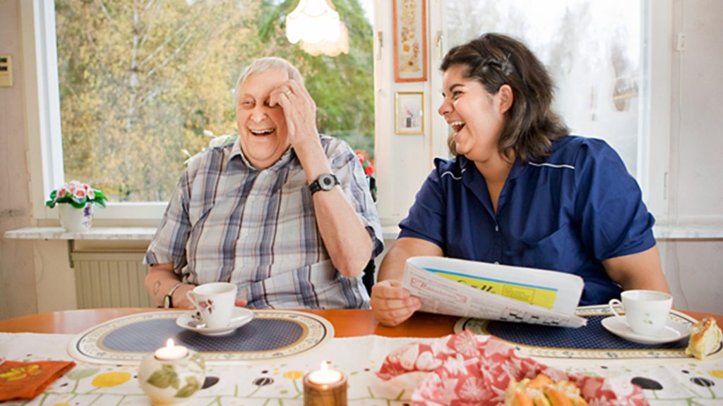 En äldre man tillsammans med ett vårdbiträde sitter och fikar