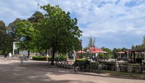 En cafébyggnad och en uteplats med bord, stolar och parasoller. Runt flera bord sitter personer och fikar. Två personer promenerar förbi den vita utescenen.