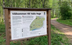 En skylt med en karta över området Valls Hage. Det står ”Välkommen till Valls Hage”. I bakgrunden syns grönskande träd och ett tillgänglighetsanpassat promenadstråk. 