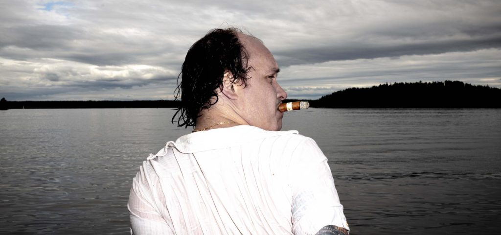 Man i halvfigur i vit t-shirt sitter vid en sjö. Himlen är grå och molnig. Mannen syns i profil, han tittar till höger.