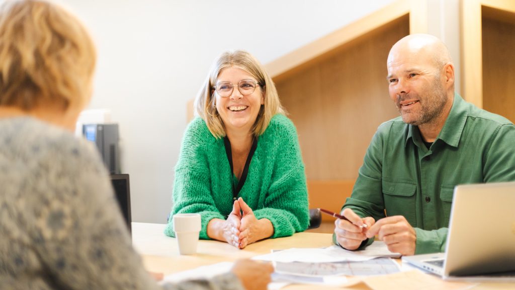 tre personer sitter vid ett bord. En man och en kvinna är synliga framifrån. En laptop står på bordet.