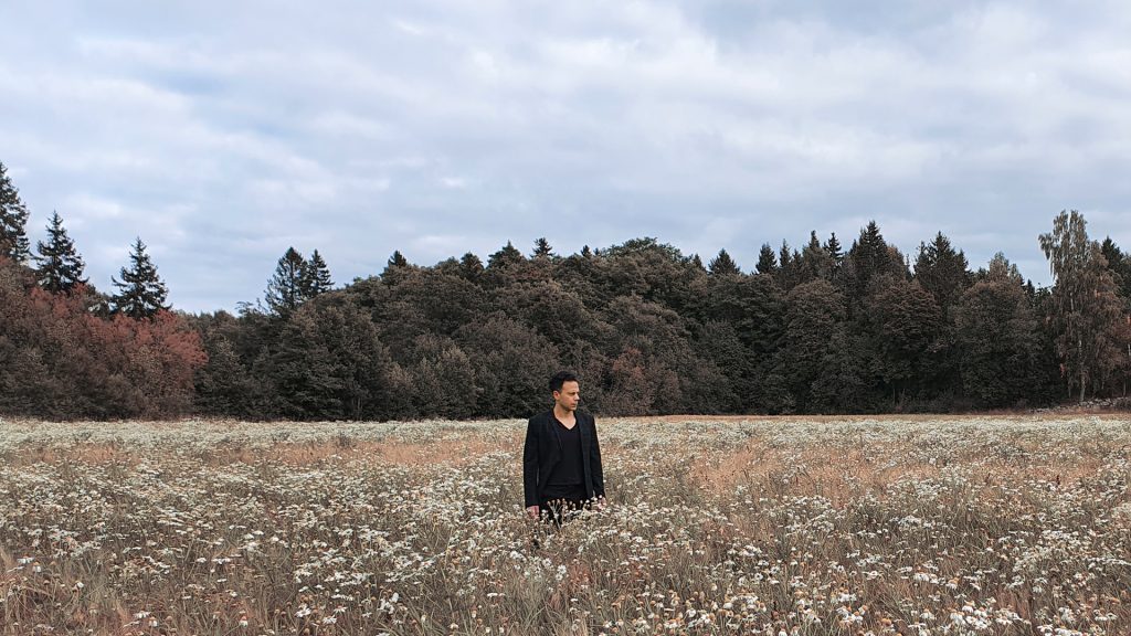 Fotografi på en person med svarta kläder och svart, kortklippt hår som står bland blommor på en häng. I bagkrunden syns en mörk skog och en molning, regntung himmel.