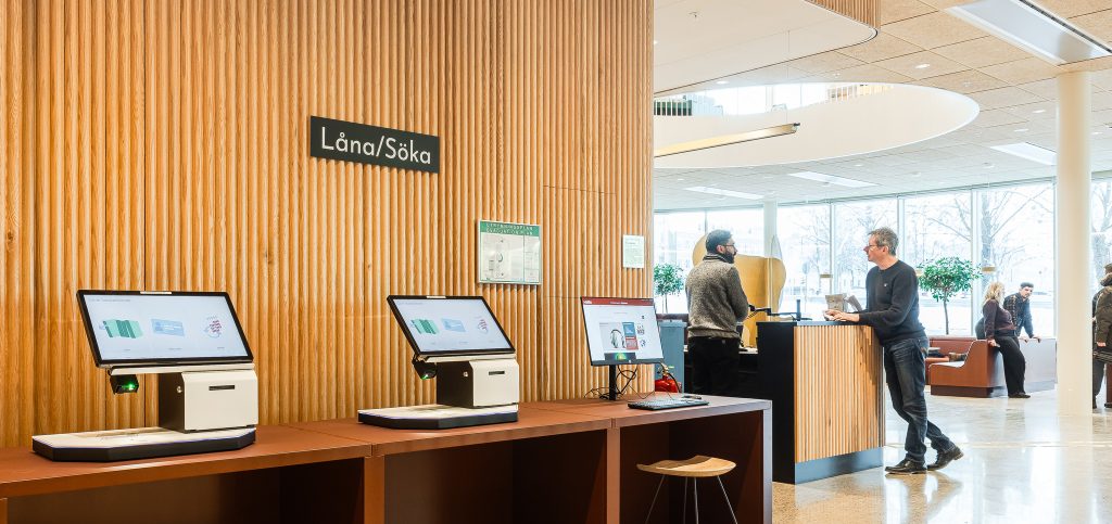 Tre skärmar som står längs en vägg på ett bibliotek. Över skärmarna står texten "sök och låna".