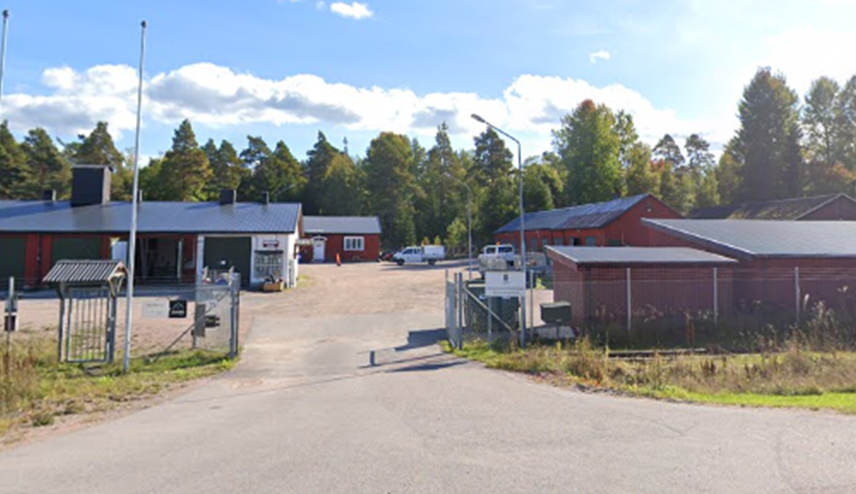 Ingång till ett område med flera röda byggnader, inhägnat med staket och omgiven av skog.