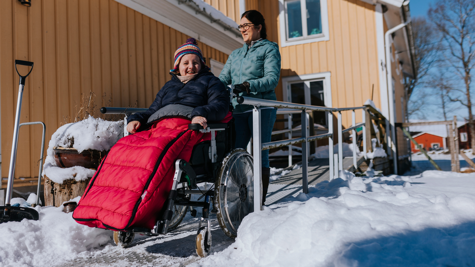 Kvinna leder ut ett barn i rullstol på en ramp i vinterlandskap