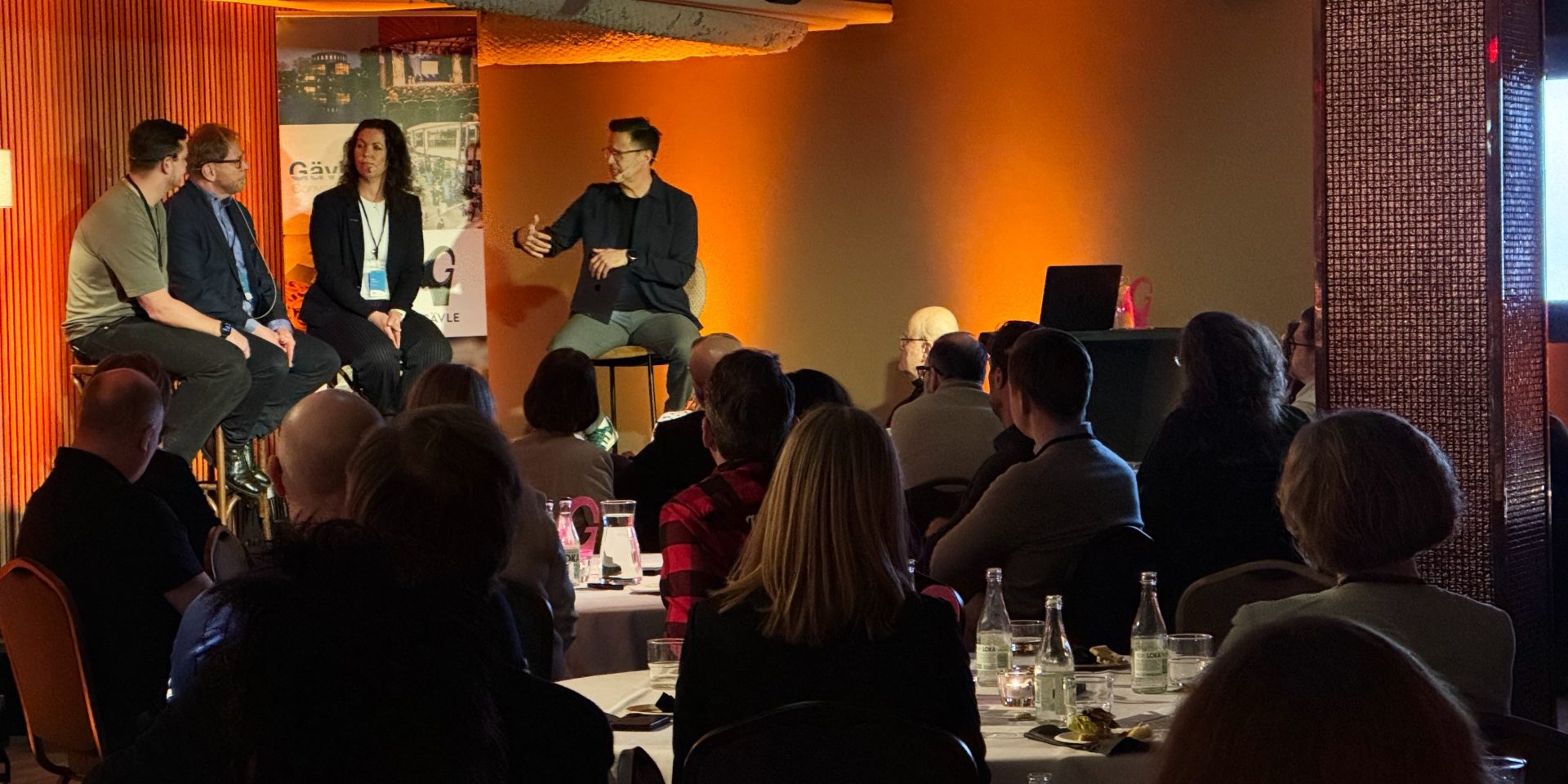 Personer som sitter på en scen och pratar och framför scenen sitter människor runt bord och lyssnar.