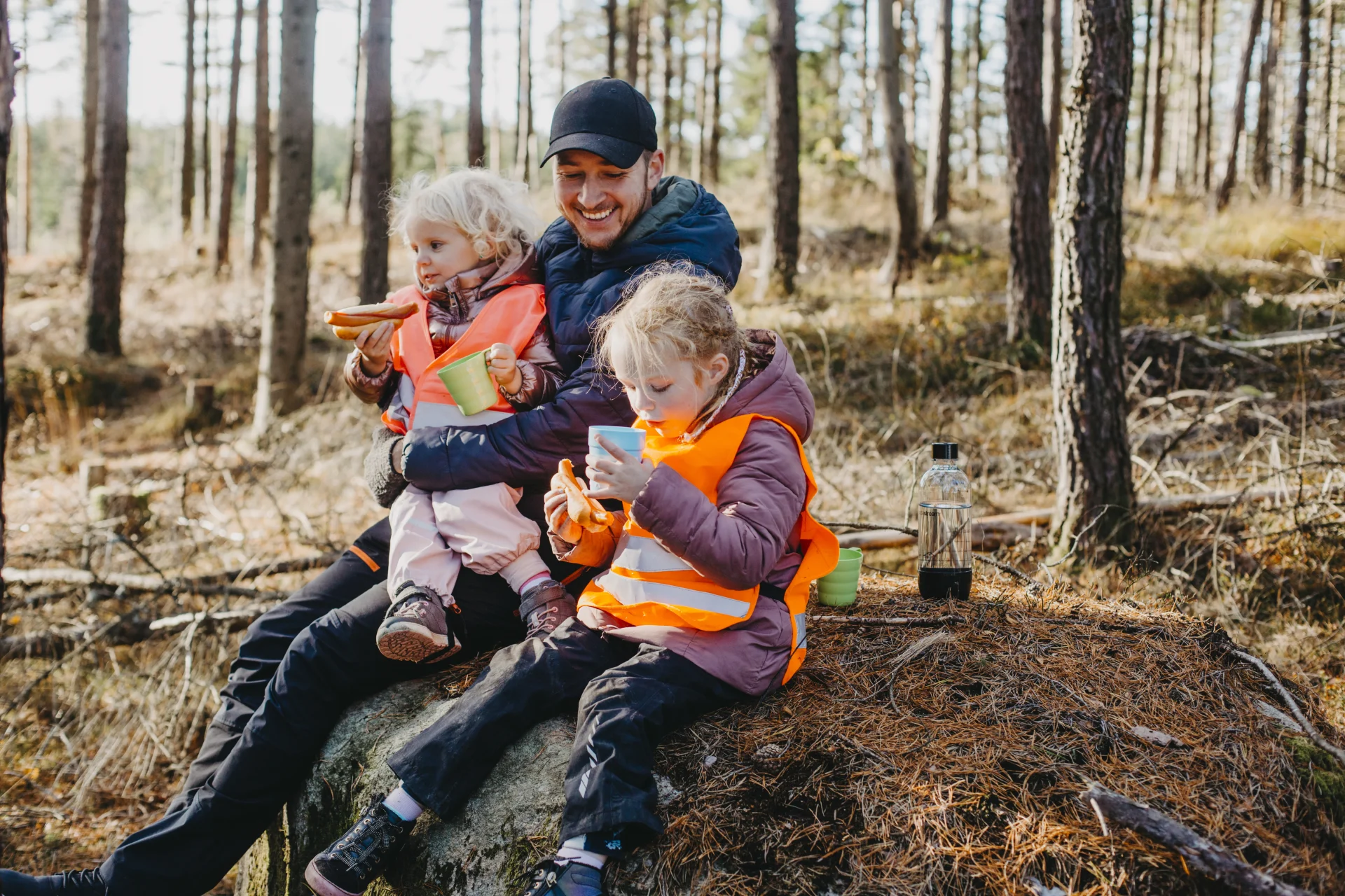 En vuxen som sitter med två barn i en skog och fikar.