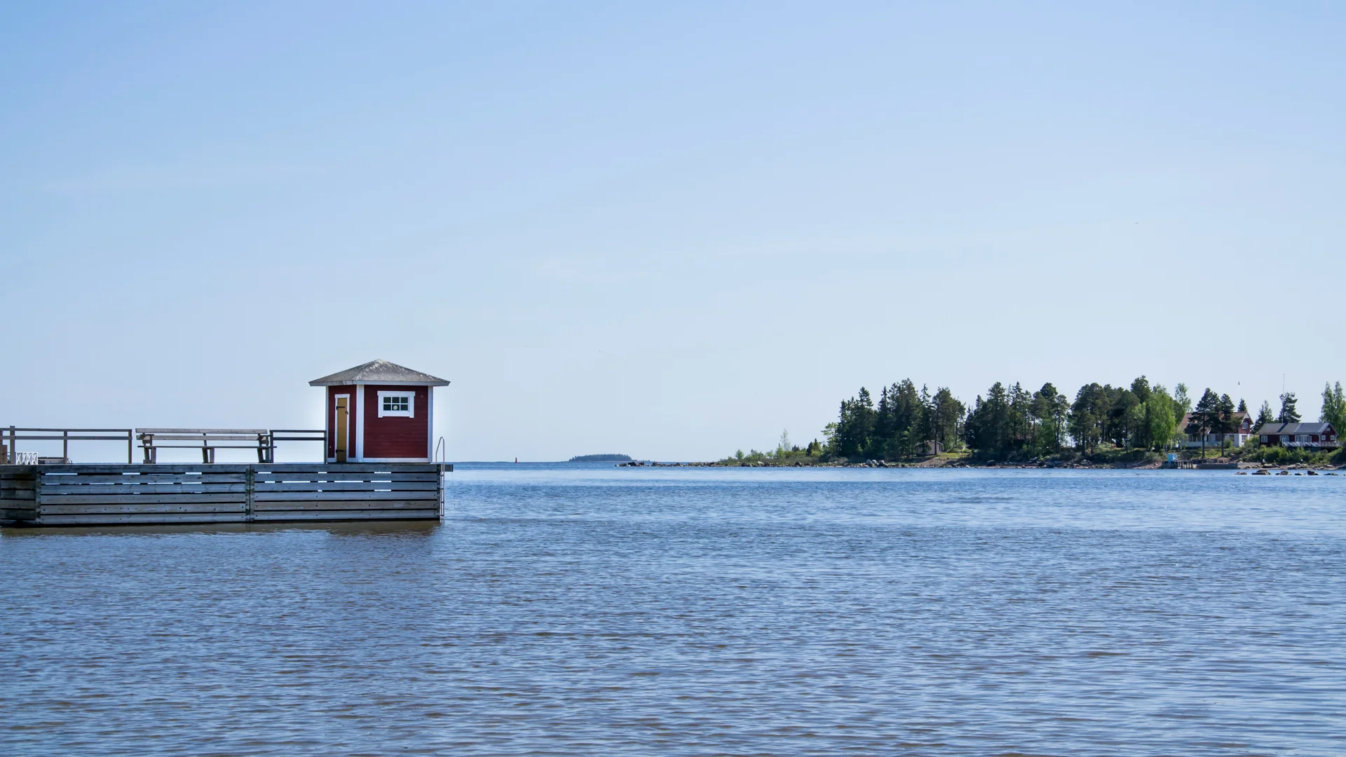 Foto av en brygga med en liten röd byggnad på, vatten och blå himmel.