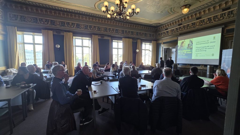 En sal med personer sittandes vid bord. En stor presentationsskärm syns i bakgrunden.